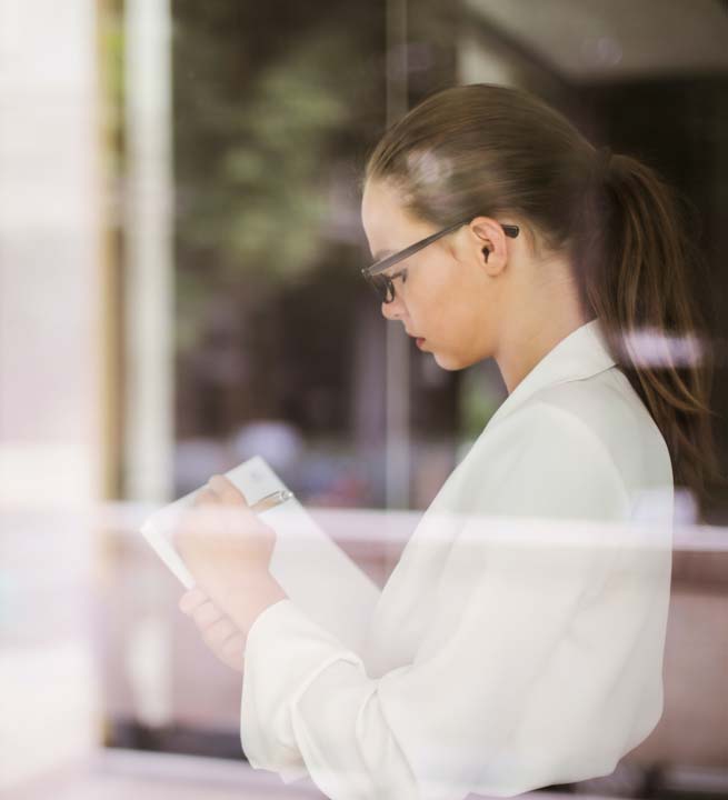 Young blonde woman with glasses and hair tied up, standing near a window, analyzing company information
