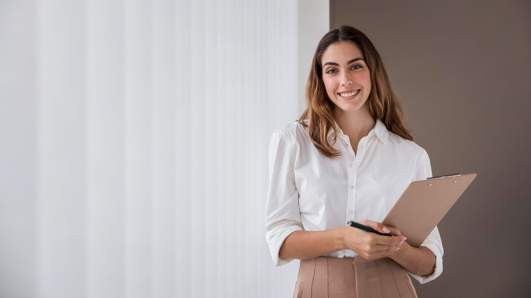 Jovem mulher de cabelos lisos, castanhos claros soltos, com blusa branca, segurando uma caneta e uma prancheta, sorrindo