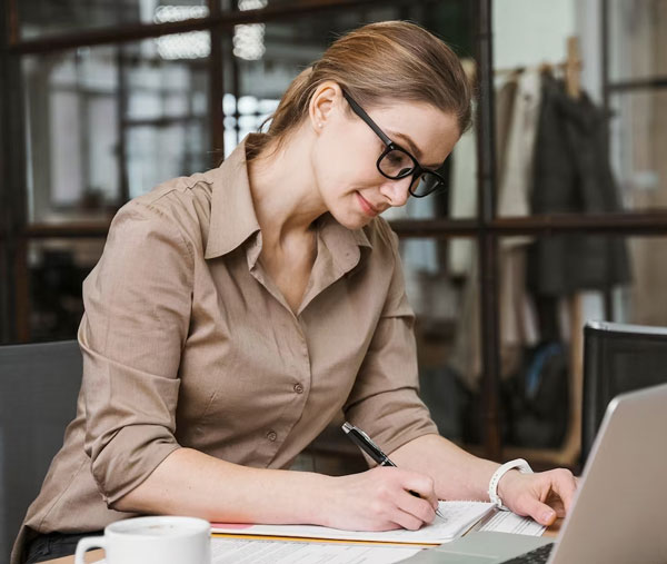 A blonde woman with glasses, her hair tied up and a beige dress shirt, taking notes