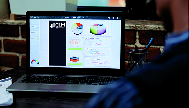 Notebook com dashboard na tela com gráficos e logo da CLM Controller em cima de mesa de escritório com porta lápis em frente de parede de tijolo